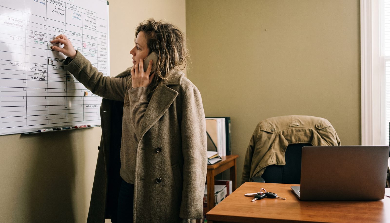 A home-care agency owner at a small office desk by a window, rota sheets and a worn diary beside the phone