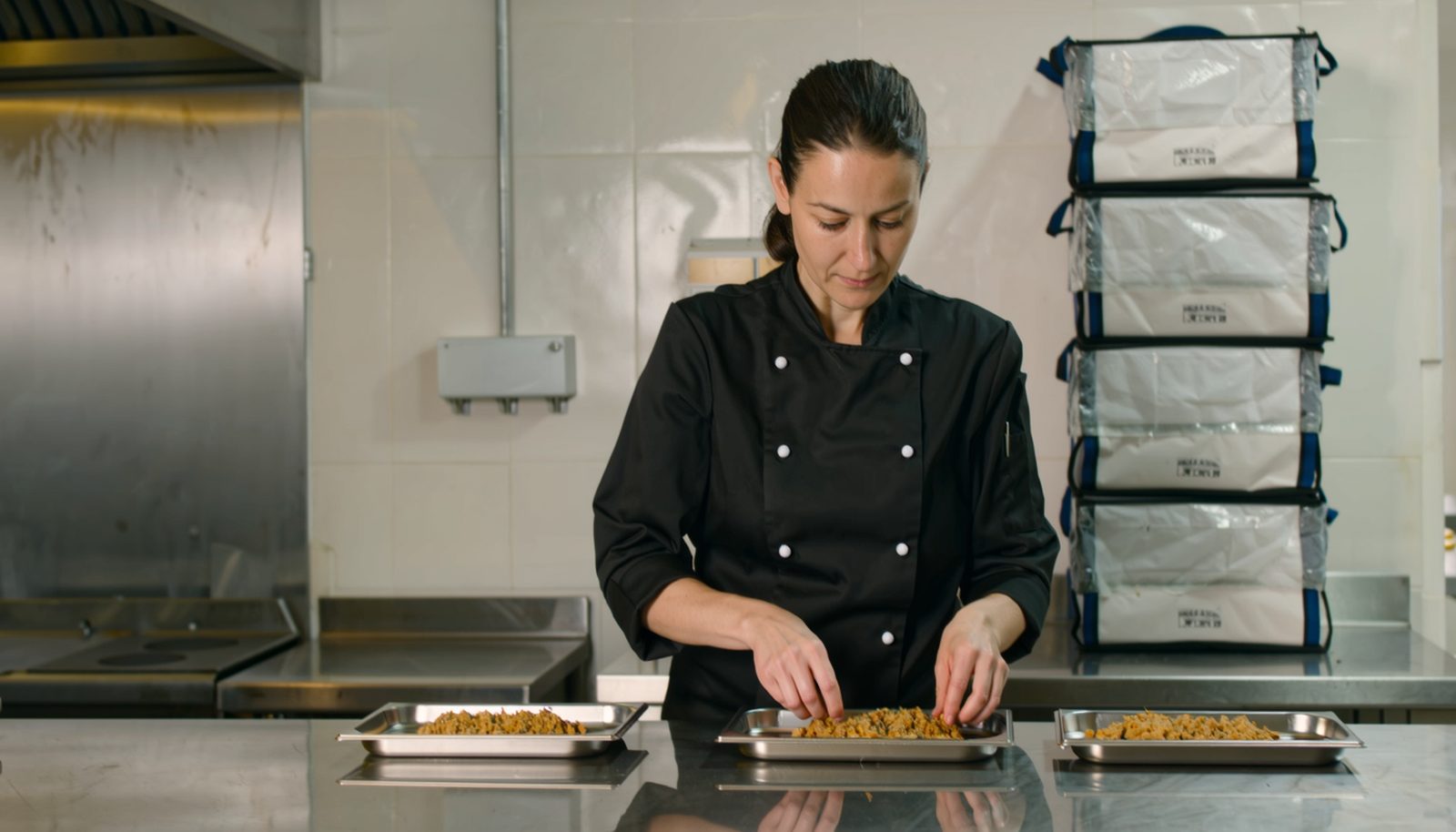 En cateringägare i svart kockjacka som lägger upp lika stora portioner på brickor för en lunchleverans till ett företag, med staplade varmhållningsboxar bakom sig