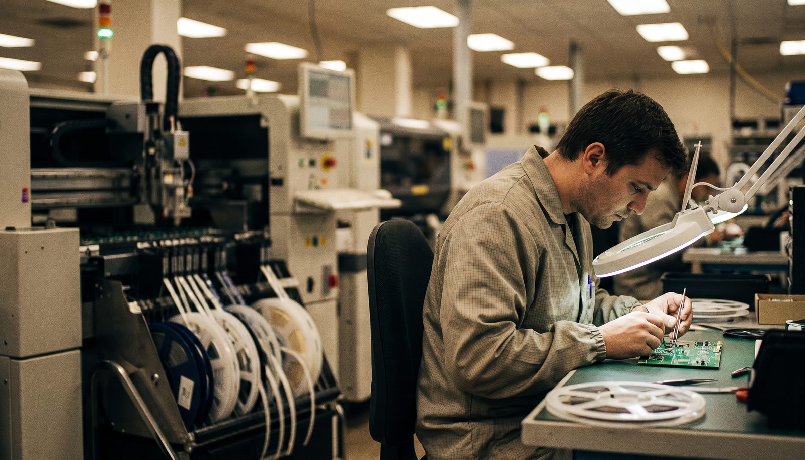 An assembler at a bench in a small electronics workshop, inspecting a populated circuit board under a magnifier lamp
