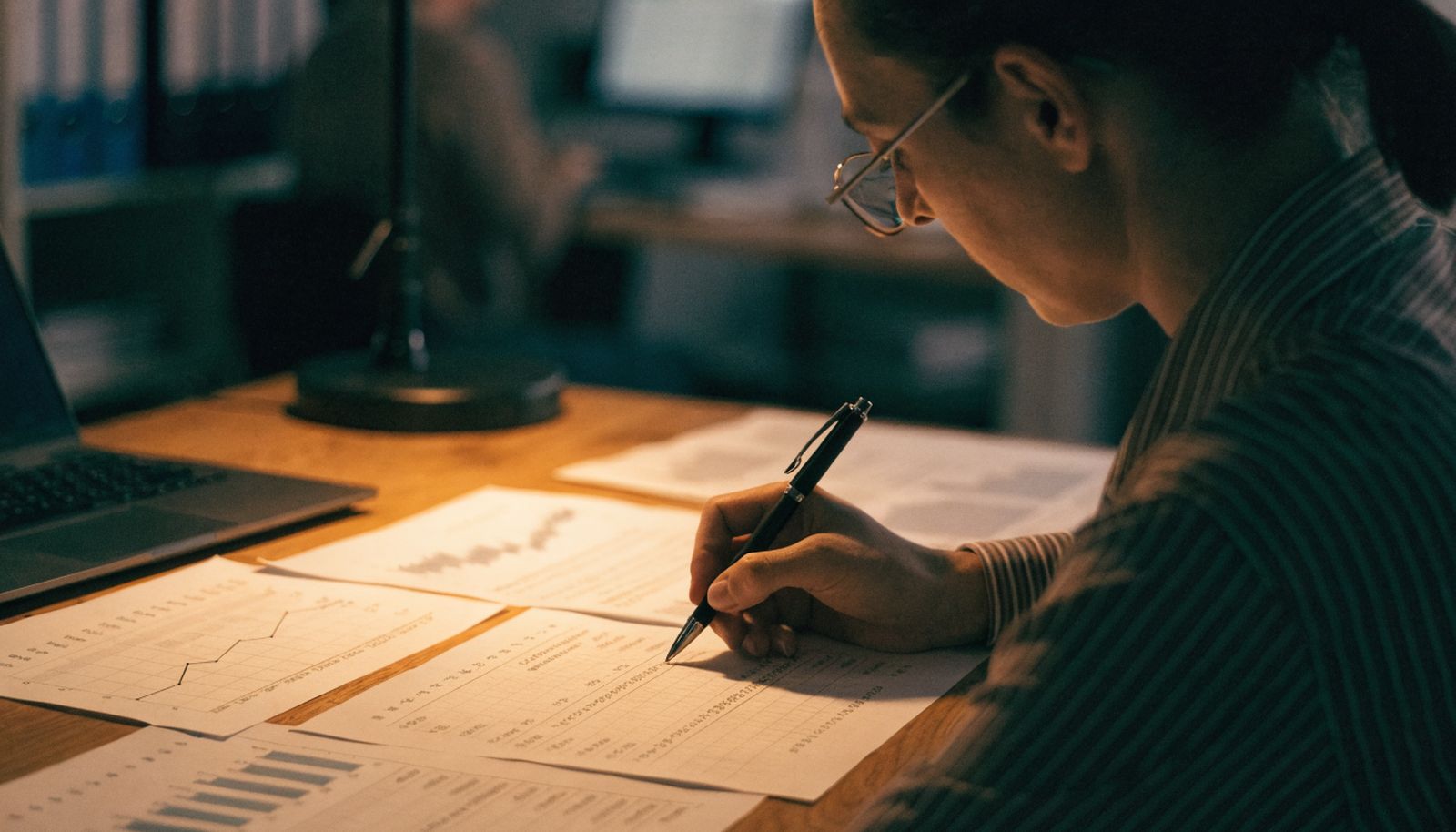 A finance consultant at a desk leafing through printed charts beside a laptop in a small office