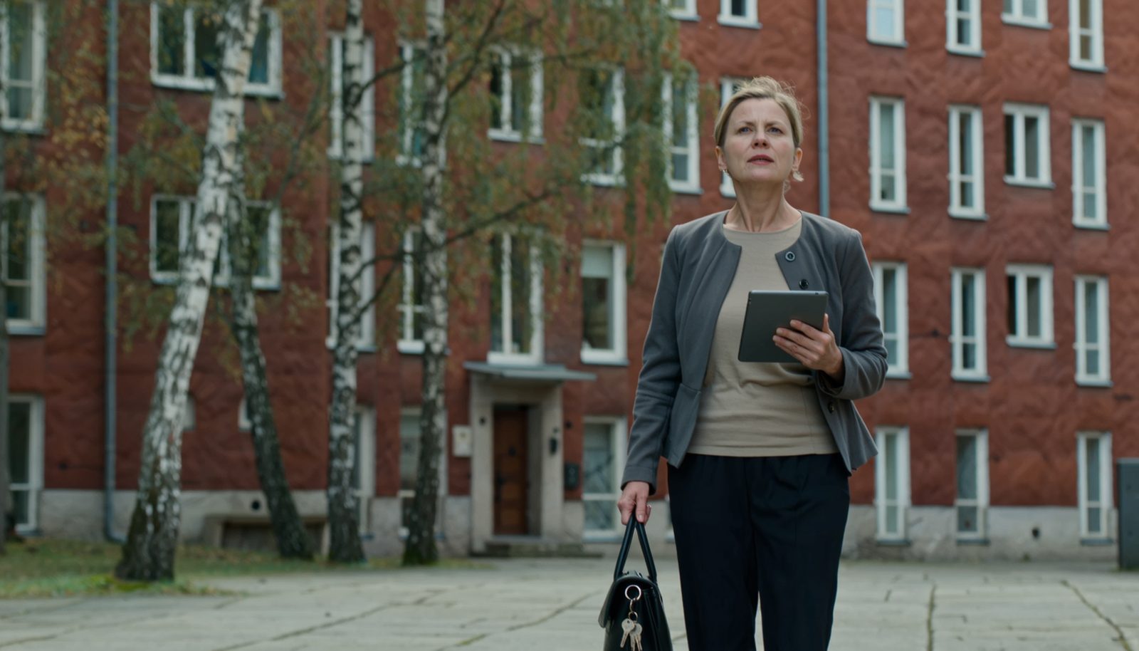 A property manager in a smart casual jacket standing in the courtyard of a 1960s residential apartment block, holding a tablet and looking up at the facade